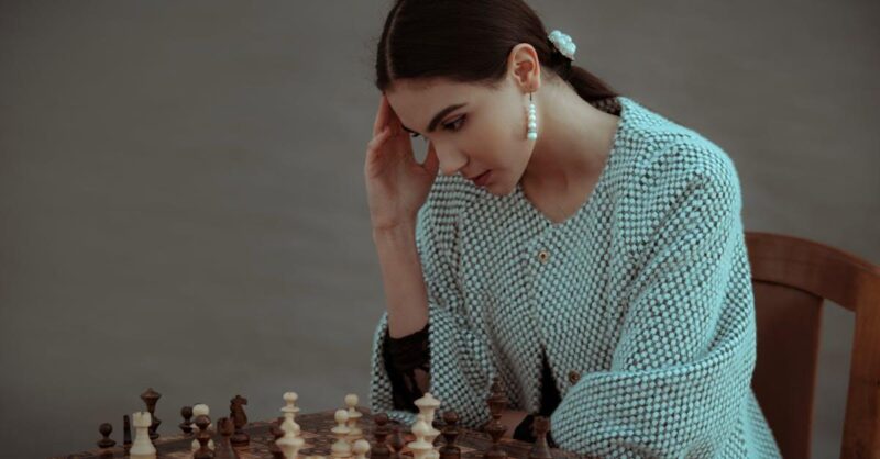 Concentrated ethnic female sitting on chair and touching head while playing chess against blurred gray background